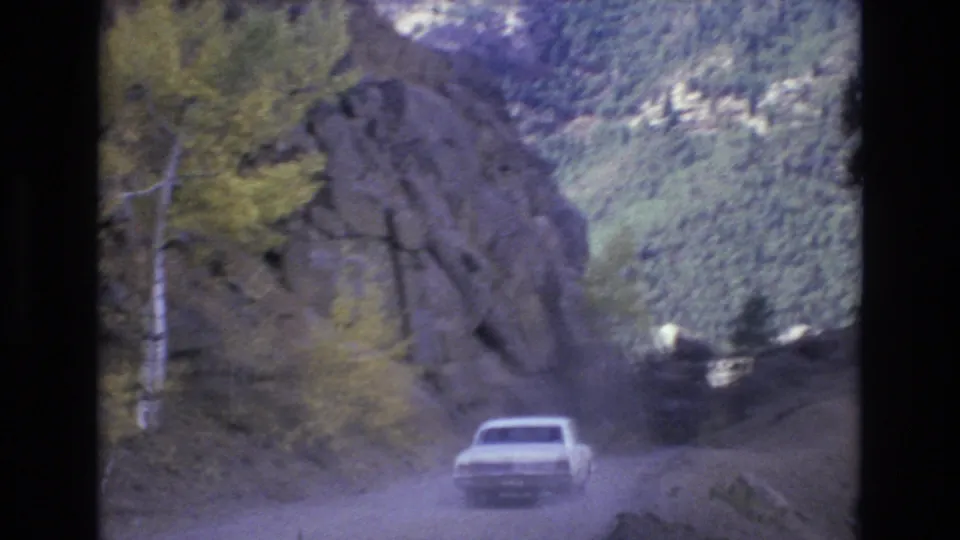 The car continues along a narrow unpaved road in the Colorado Springs mountain landscape, with rock walls, trees, and a forested valley visible ahead.