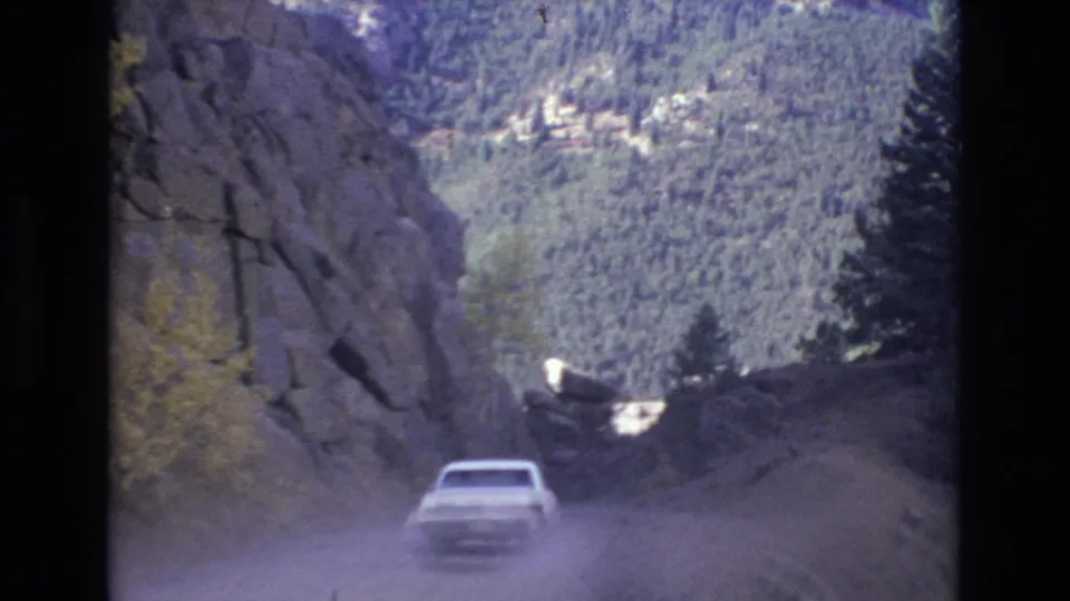 A rear view of the white car shows it descending or curving along a dusty mountain road, with steep rock, trees, and a broad wooded slope in the distance.