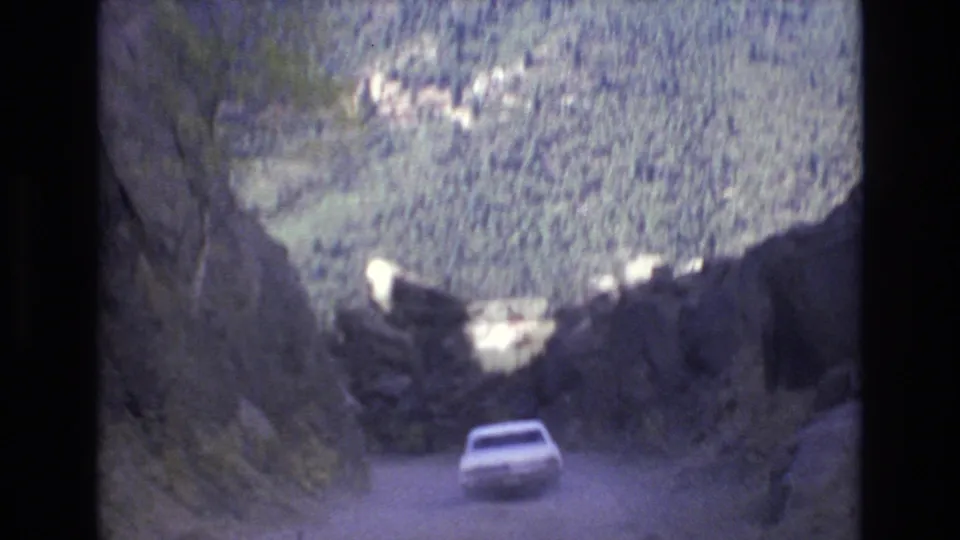 The car moves farther along the winding dirt road through the Colorado Springs mountain valley, framed by dark rock walls and a wooded hillside.
