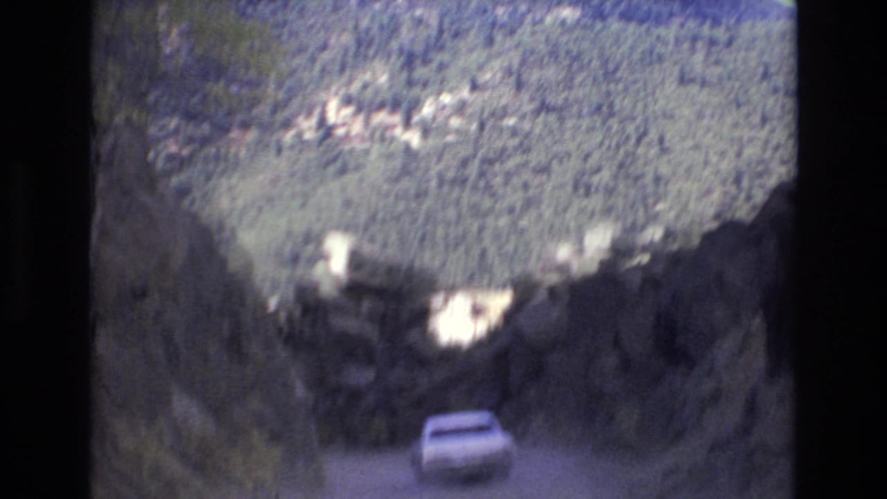 1973 Colorado Springs home-movie frame showing a white automobile on a steep unpaved mountain road bordered by rugged rocks and distant forested slopes.