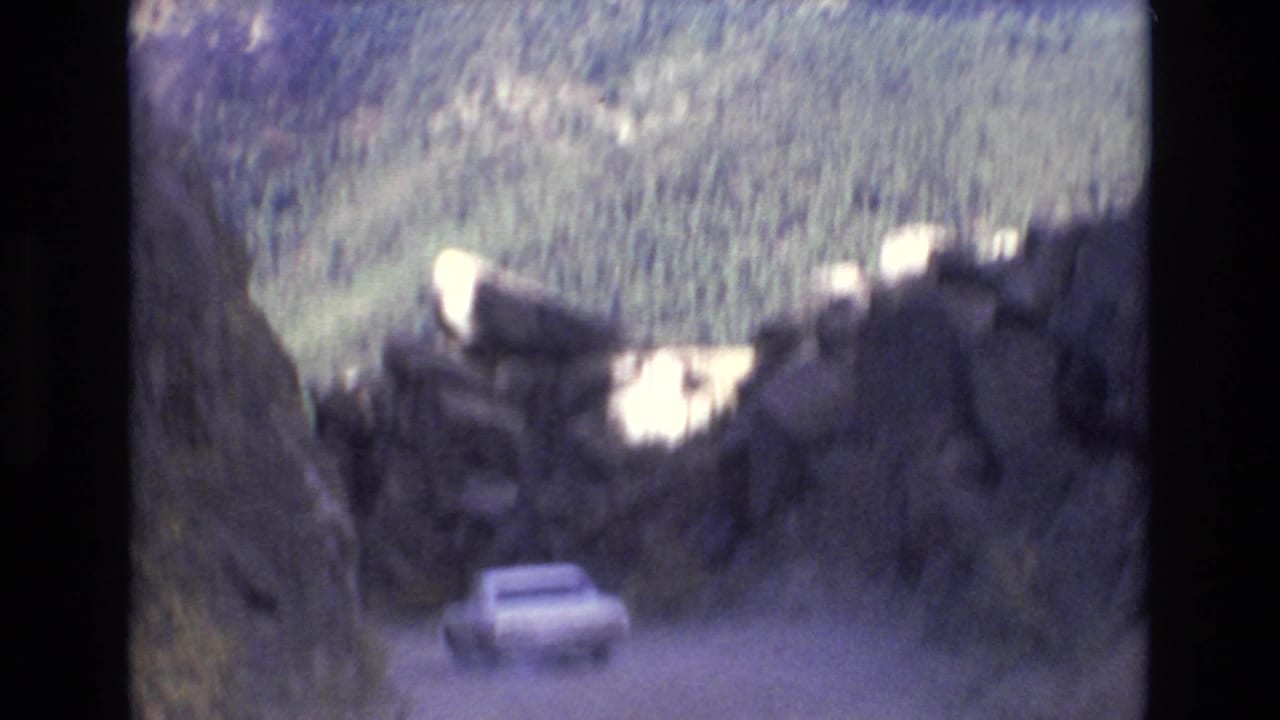 White car farther down a dusty mountain road framed by rocky outcrops and tree-covered hills.