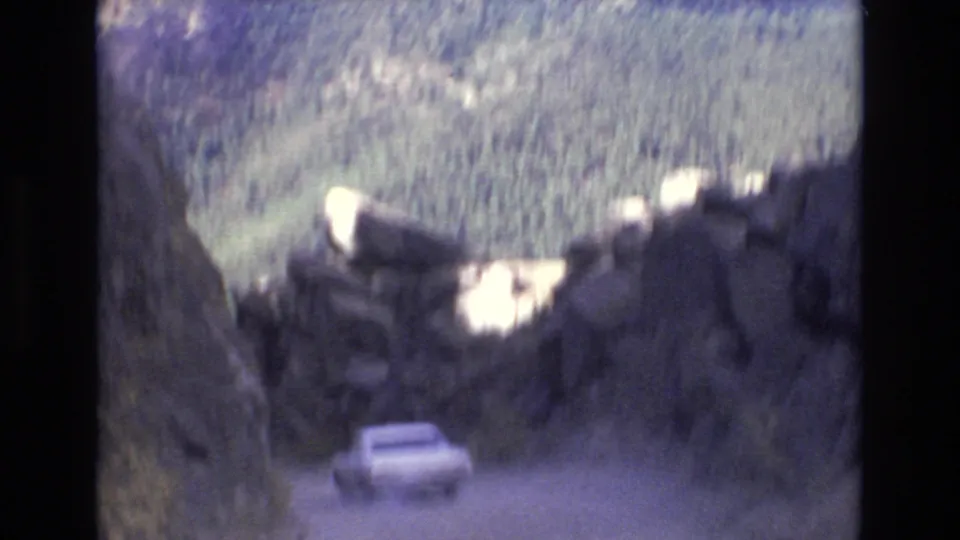White automobile continues downhill through a narrow rocky passage in Colorado Springs, with sunlit boulders and forested mountain terrain behind it.