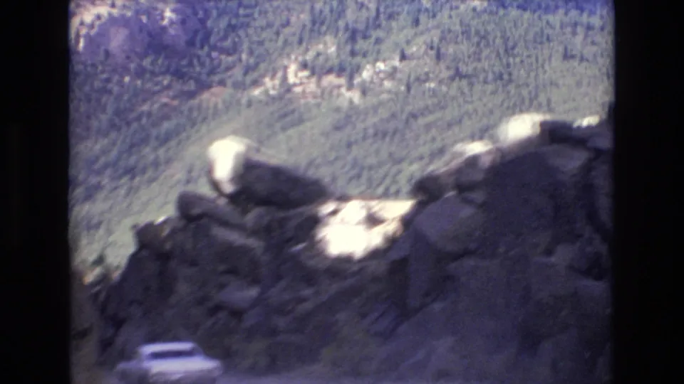 Home-movie view of rugged Colorado Springs rock formations above a steep road, with the white car partly visible below and wooded hills in the background.