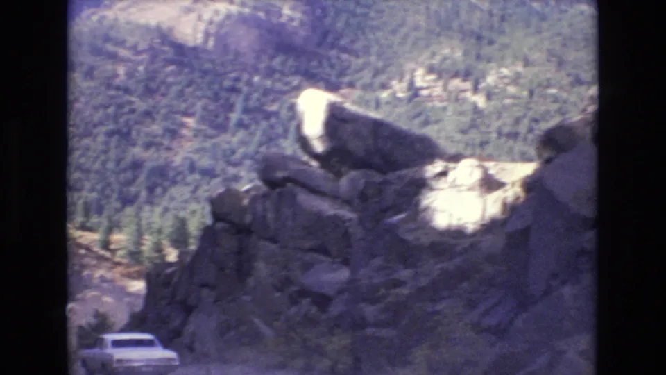 A white automobile appears below rugged boulders on a steep mountain road in the Colorado Springs 1973 home-movie footage, with dense trees across the slope behind.