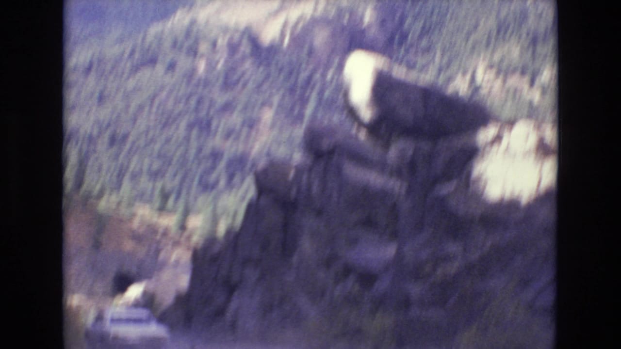 A white car drives along a winding mountain road near Colorado Springs, with rugged rock outcrops and pine-covered hills visible in soft 1970s 8mm color.