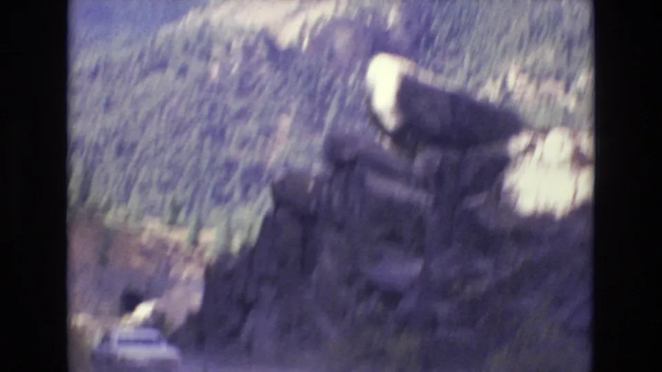 A white car drives along a winding mountain road near Colorado Springs, with rugged rock outcrops and pine-covered hills visible in soft 1970s 8mm color.