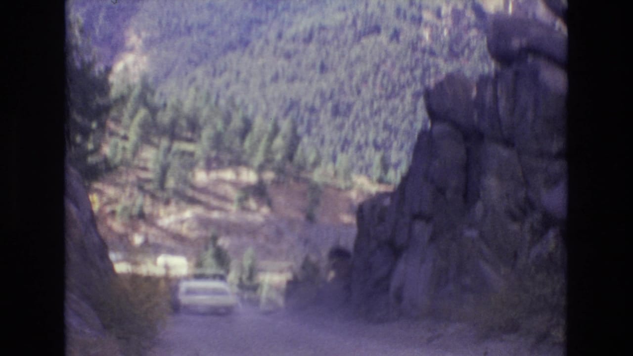 The white sedan moves away along a mountain road bordered by rock walls and pine forest in archival 1973 Colorado Springs-area footage.