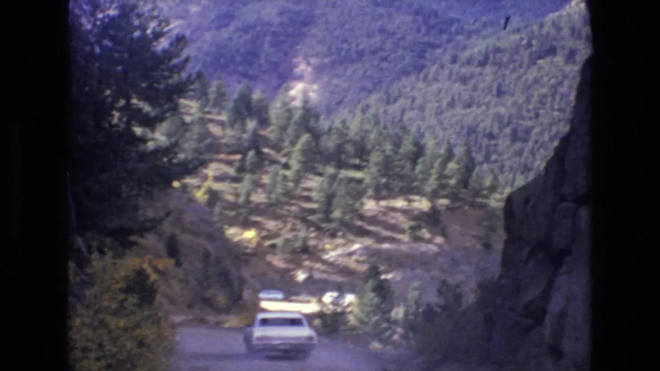 A white car travels downhill on a curving mountain road, with pine trees, rocky terrain, and distant slopes visible in the Colorado Springs travel scene.