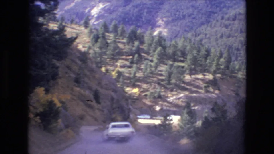 The white car continues along the dusty mountain road, with forested hills, rock faces, and small roadside vehicles or structures in the background.
