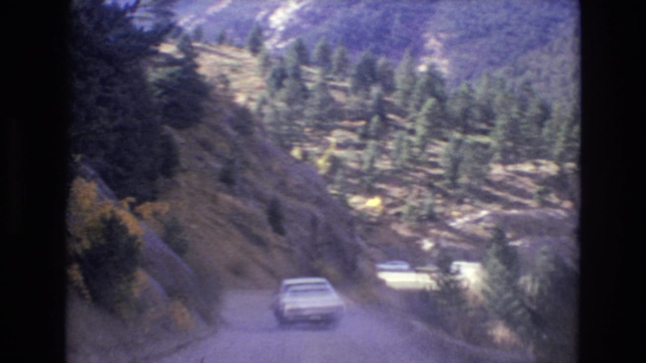 1973 Colorado Springs home-movie frame showing a white car ahead on a winding dusty mountain road with pine trees and rocky hillsides.