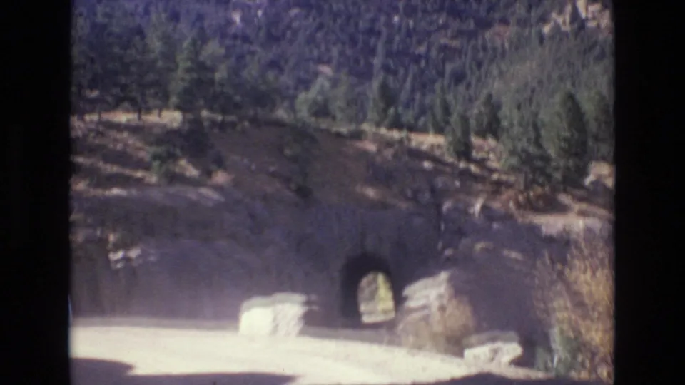 1973 Colorado Springs 8mm frame showing a small tunnel opening in a rocky mountainside beside a bright road and pine-covered terrain.