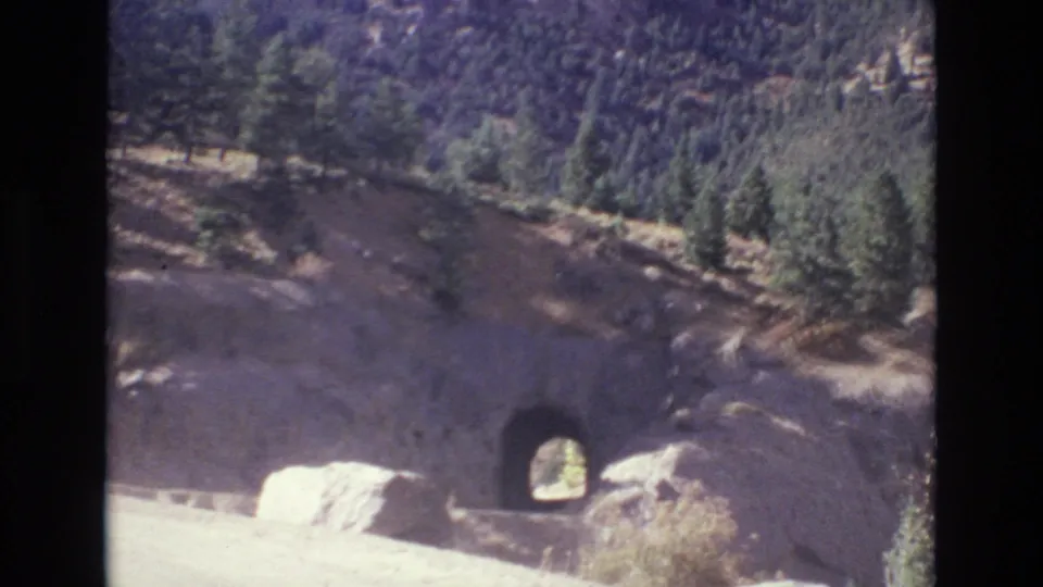 1973 Colorado Springs home-movie frame showing a winding mountain road passing a rock-cut tunnel with pine forest and rugged slopes behind it.