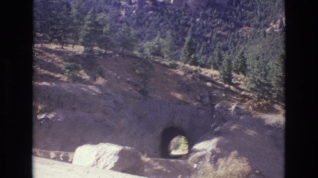 1973 Colorado Springs-area 8mm frame showing a rocky mountain slope, evergreen trees, and a dark tunnel opening along a roadside.