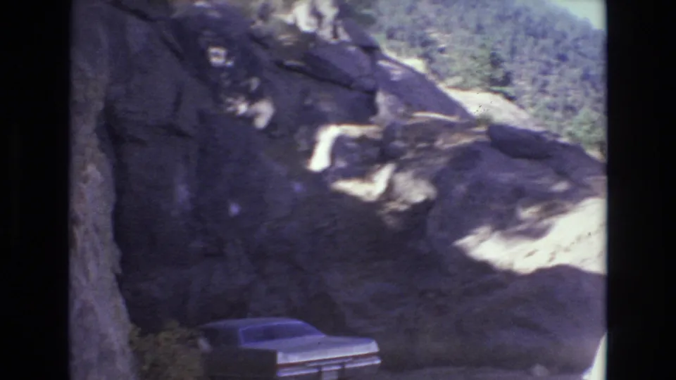 Handheld 8mm frame from the mountain road sequence showing a dark rock face, bright exposed stone, and the rear portion of a car near the lower edge.