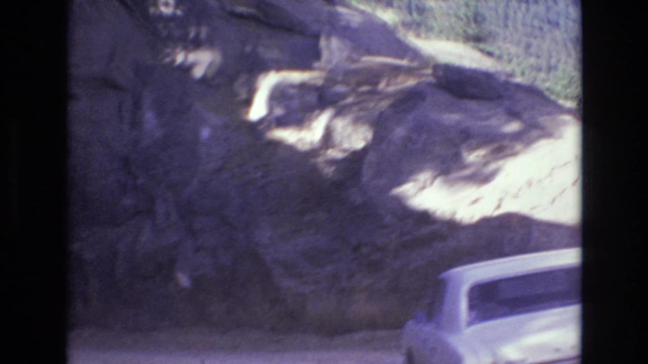 A pale vintage car appears near a rocky tunnel entrance during a 1973 home-movie road sequence in Colorado Springs, with soft color and handheld framing.