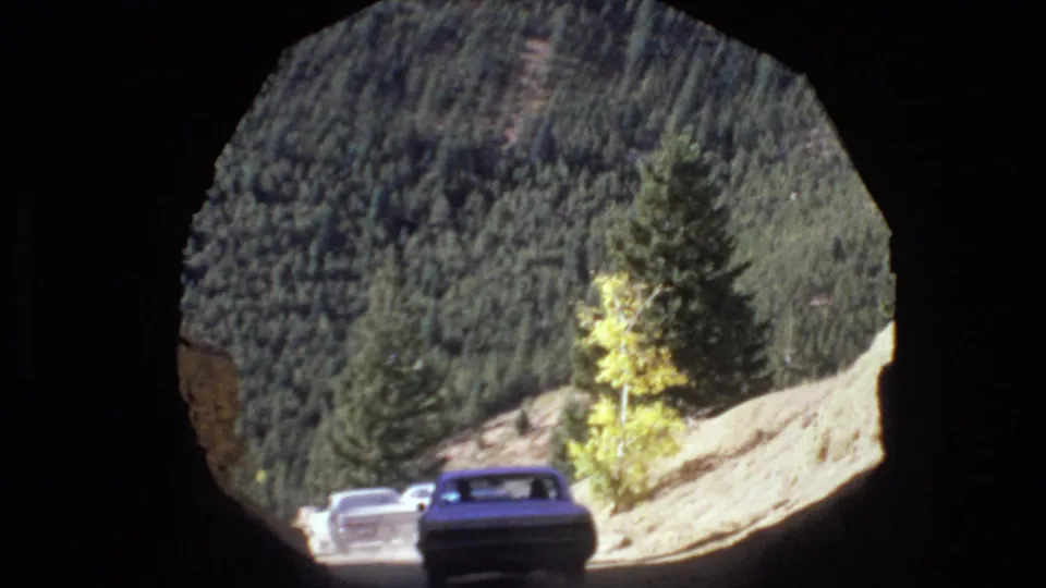 From inside the tunnel, two older cars are visible on the mountain road beyond, with evergreen-covered slopes and a yellow-leaved tree in daylight.