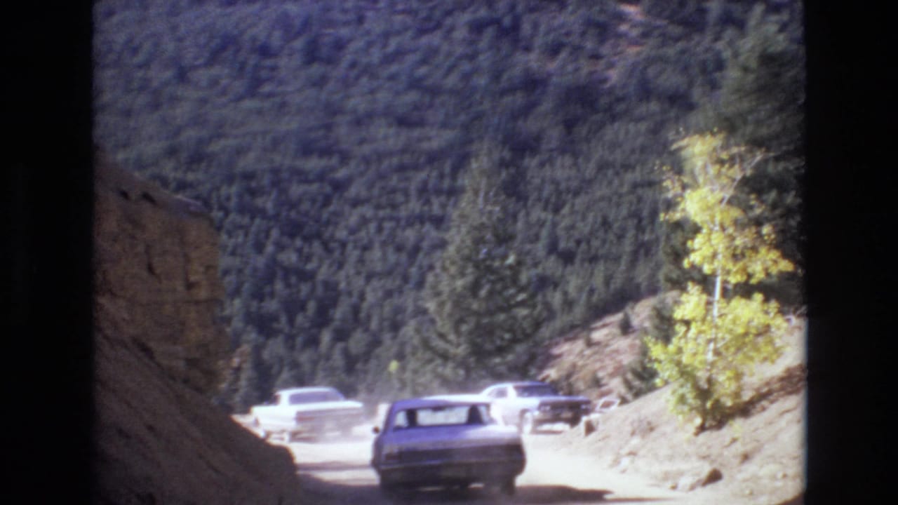 Several cars travel on an unpaved mountain road near Colorado Springs, Colorado, in 1973, with evergreen slopes and roadside rock visible in soft 8mm color footage.
