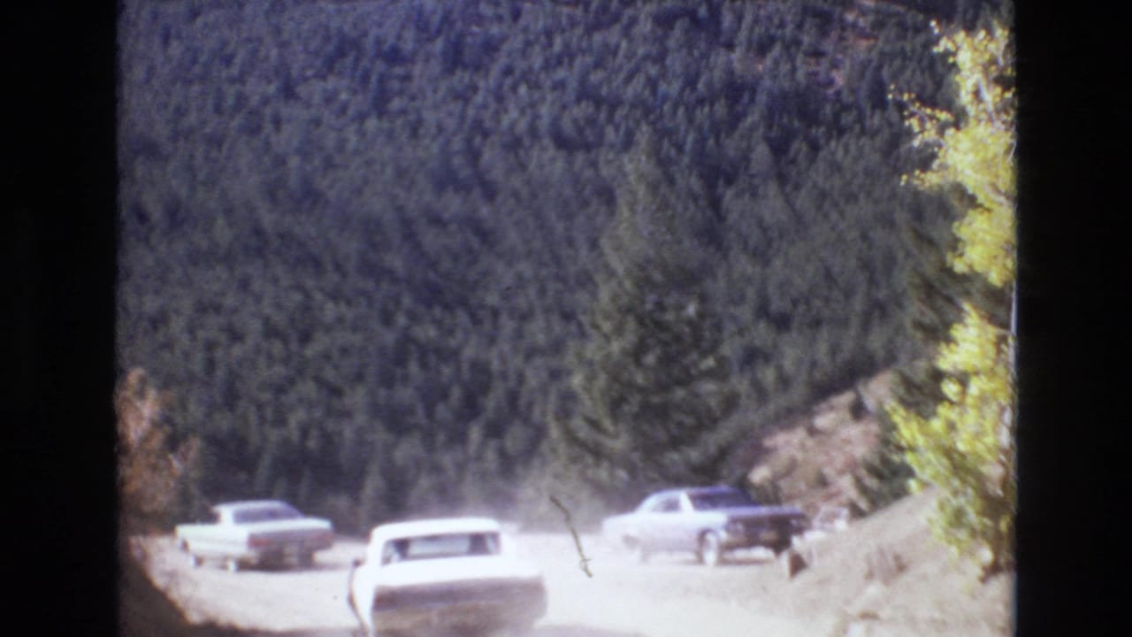 Light-colored cars continue along a dusty mountain road in the Colorado Springs area, with dense evergreen trees and sun glare softening the 8mm frame.