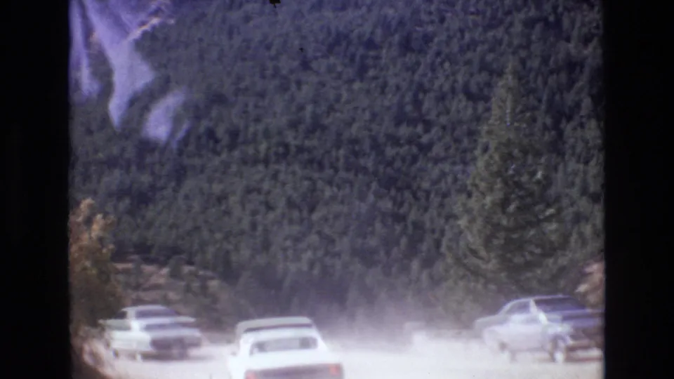 Vehicles on an unpaved Colorado mountain road raise dust against a backdrop of dark evergreen trees and rocky roadside terrain in 1973 8mm home-movie footage.