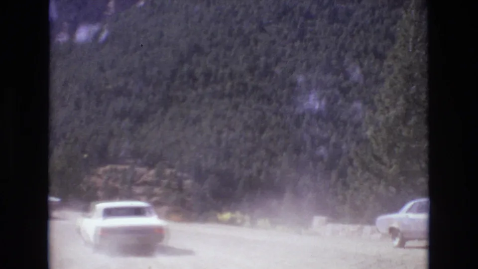 A car moves away through lingering dust on a sunlit unpaved mountain road, with forested slopes and another vehicle partly visible at the right edge.