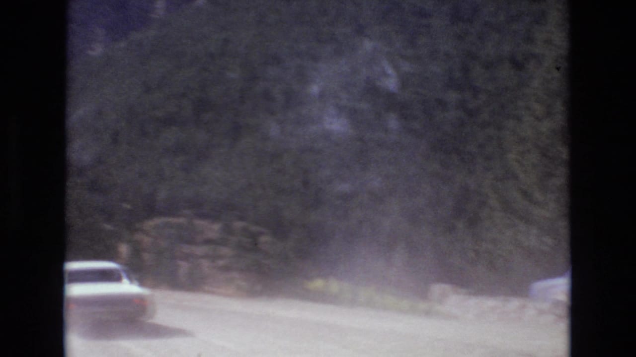 White car driving on a dusty dirt road through a wooded mountain area.