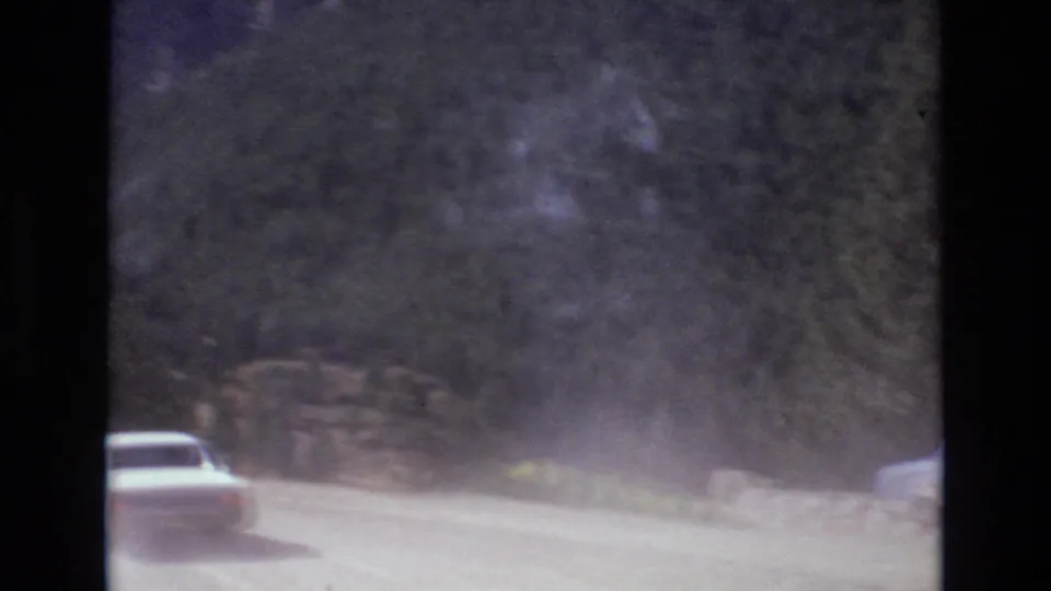 A white sedan travels along an unpaved wooded mountain road near Colorado Springs, with dust trailing behind in 1973 8mm home-movie footage.