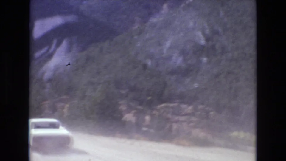 A car moves away on a curved unpaved road through a wooded mountain setting, with soft 8mm grain and dust visible in the air.