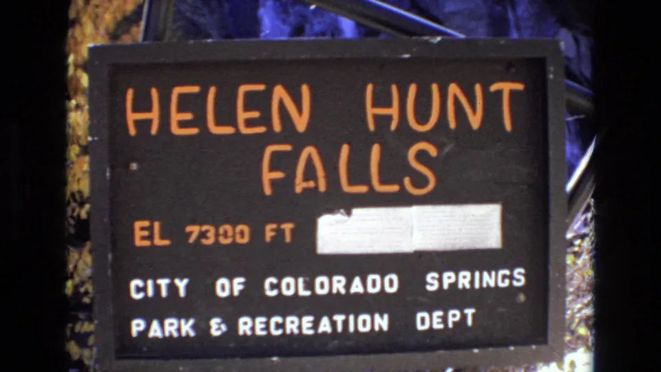 1973 8mm frame showing a lower handheld view of the Helen Hunt Falls sign in Colorado Springs, including the elevation line, patched rectangle, and municipal park text.