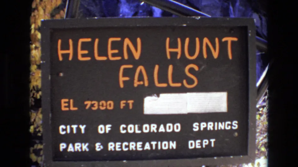 Final 1973 home-movie frame of the Colorado Springs Helen Hunt Falls sign, with the falls name, 7300 ft elevation, municipal park lettering, and surrounding dark outdoor background visible.