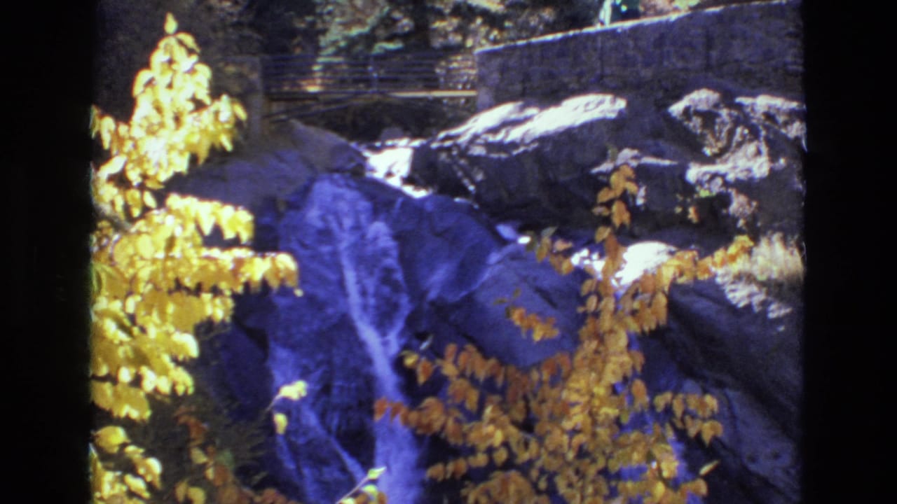 1973 Colorado Springs 8mm frame showing a small waterfall among dark rocks, bright yellow autumn leaves, trees, and a stone wall with railing in the background.