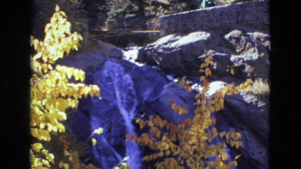1973 Colorado Springs 8mm frame showing a small waterfall among dark rocks, bright yellow autumn leaves, trees, and a stone wall with railing in the background.
