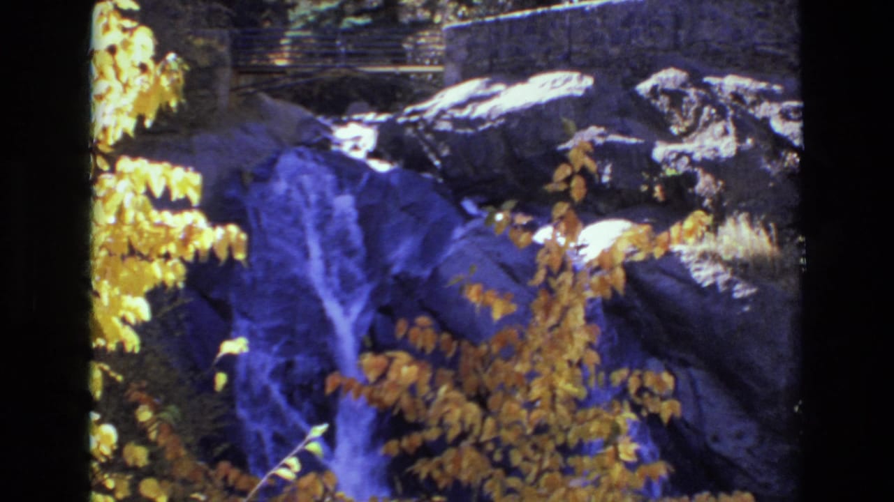 1973 Colorado Springs 8mm frame with blue-purple water cascading through shaded rocks, golden foliage in the foreground, and park-like stonework behind.