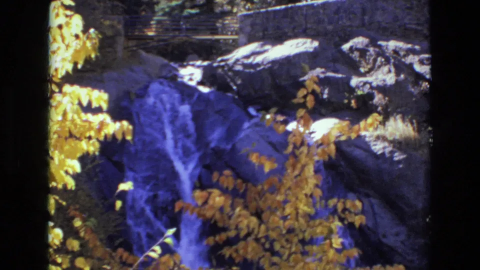 1973 Colorado Springs 8mm frame with blue-purple water cascading through shaded rocks, golden foliage in the foreground, and park-like stonework behind.