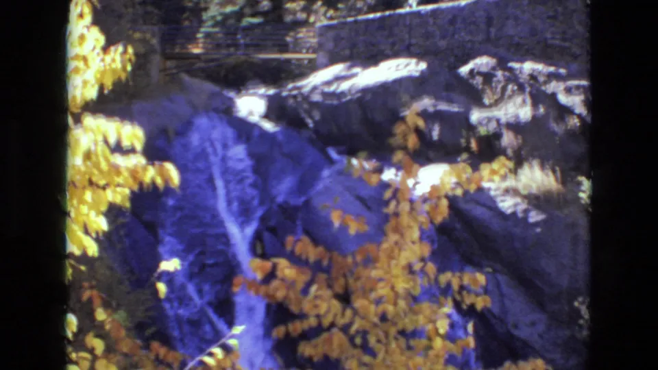 1973 Colorado Springs 8mm frame of a shaded autumn waterfall scene, with golden leaves crossing the foreground and stone park features above the rocks.