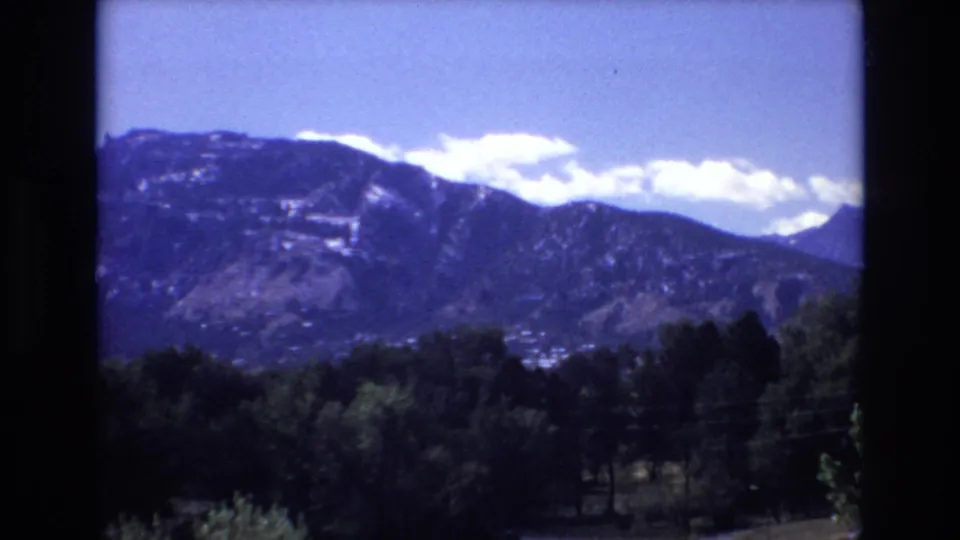 1973 Colorado Springs scenic frame showing a distant mountain ridge, bright clouds, blue sky, and trees in the lower foreground.