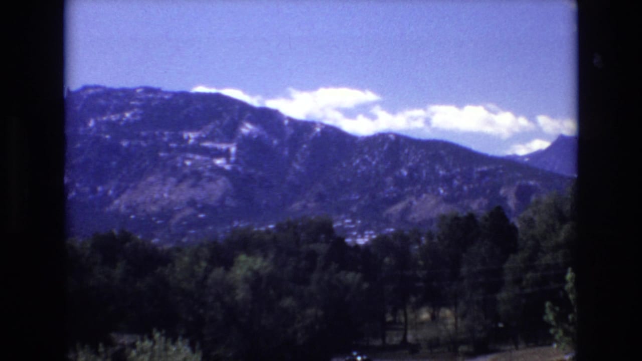 1973 Colorado Springs home-movie frame showing a mountain ridge beneath bright clouds, with green trees and dark 8mm frame borders in the foreground.