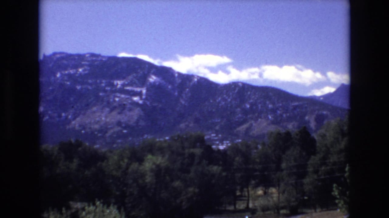 Blue sky and white clouds above a dark mountain ridge with trees below.