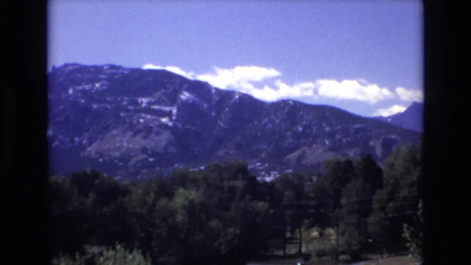 A 1973 Colorado Springs 8mm view holds on the mountain range, with bright clouds along the ridge and a shadowed tree line below.