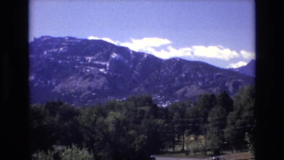 The home-movie frame shows the Colorado Springs mountain range under drifting white clouds, with dense trees and a faint roadside strip near the bottom.