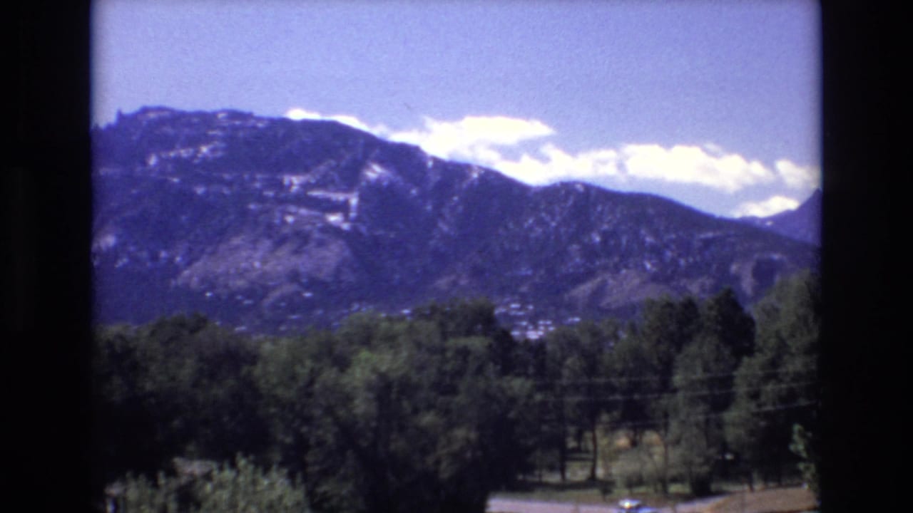 1973 Colorado Springs home-movie frame showing a snow-patched mountain range beyond dark green trees, a small distant town, and faint power lines.