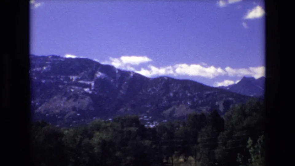 1973 Colorado Springs home-movie frame showing mountains with snow patches, a bright blue sky, white clouds, trees in shadow, and a small distant settlement.