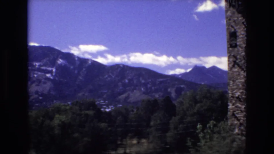 1973 Colorado Springs 8mm frame with a mountain range stretching across the background, dark trees below, scattered white clouds, distant buildings, and a dark vertical border at the right edge.