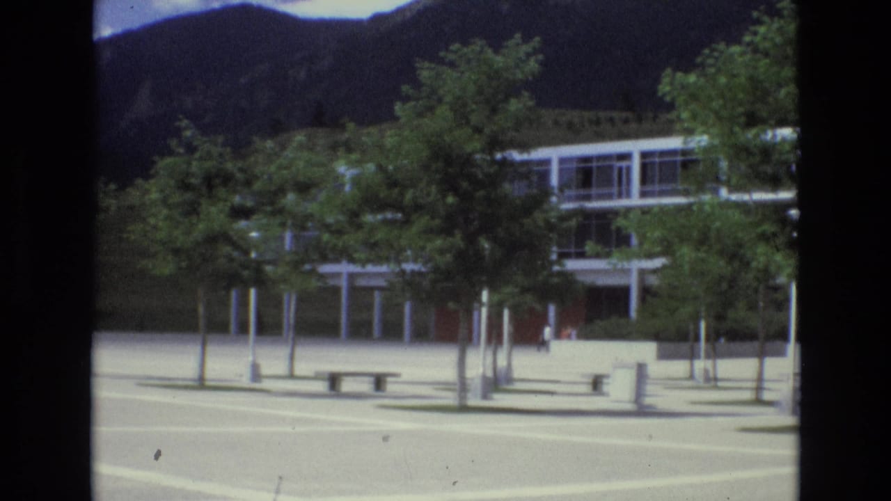 The camera view holds on the modernist facade and raised structure across a pale plaza, with planted trees partially blocking the building and mountains in the background.