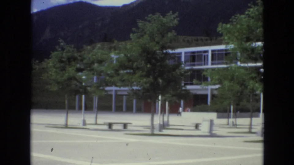 The camera view holds on the modernist facade and raised structure across a pale plaza, with planted trees partially blocking the building and mountains in the background.
