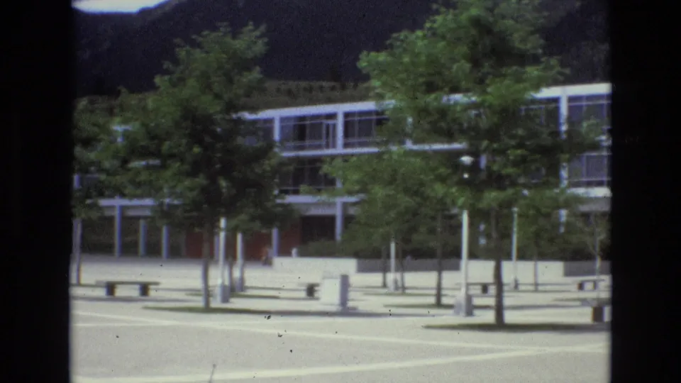 A closer panning frame shows benches, pale pavement markings, young trees, and the lower level of the modern glass building in Colorado Springs.