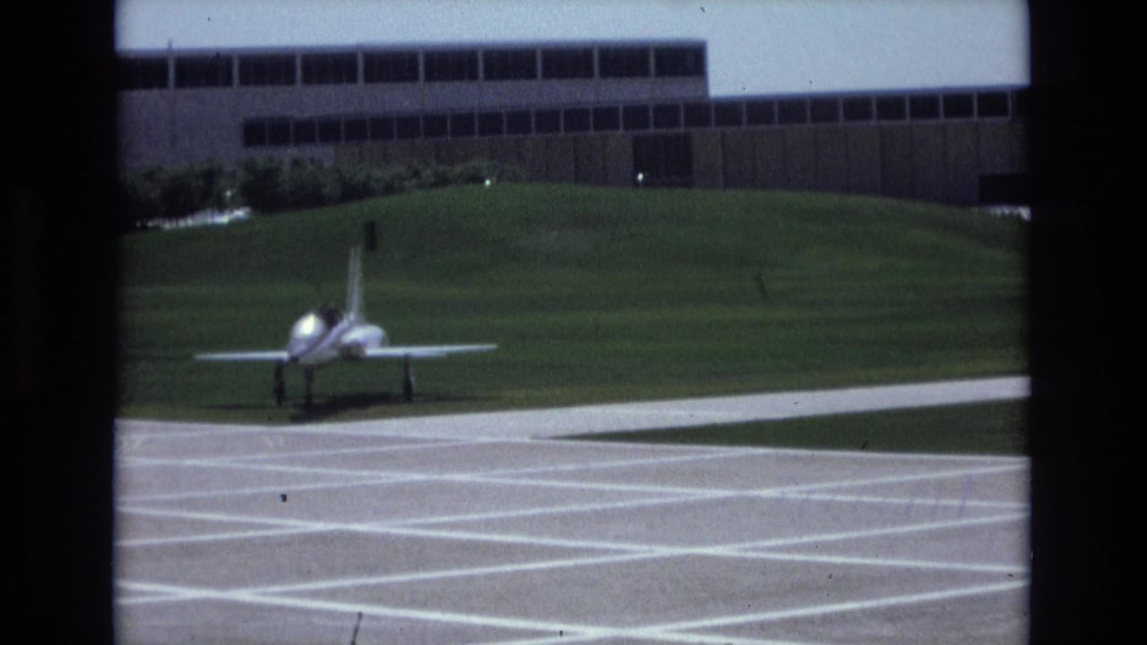 1973 Colorado Springs 8mm frame showing a small military jet parked or taxiing on a marked paved area near grass and a large modern building.