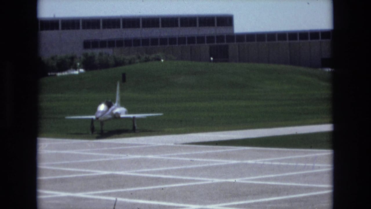 1973 Colorado Springs 8mm frame with a distant military jet centered beyond white pavement markings, grass, and a large institutional building.