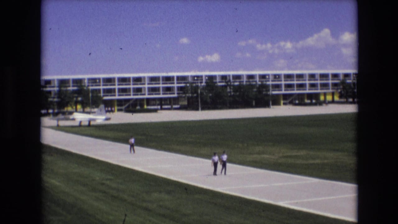 1973 Colorado Springs 8mm frame showing two distant men on a pale concrete walkway, with green grass, a long modern building, and blue sky behind them.