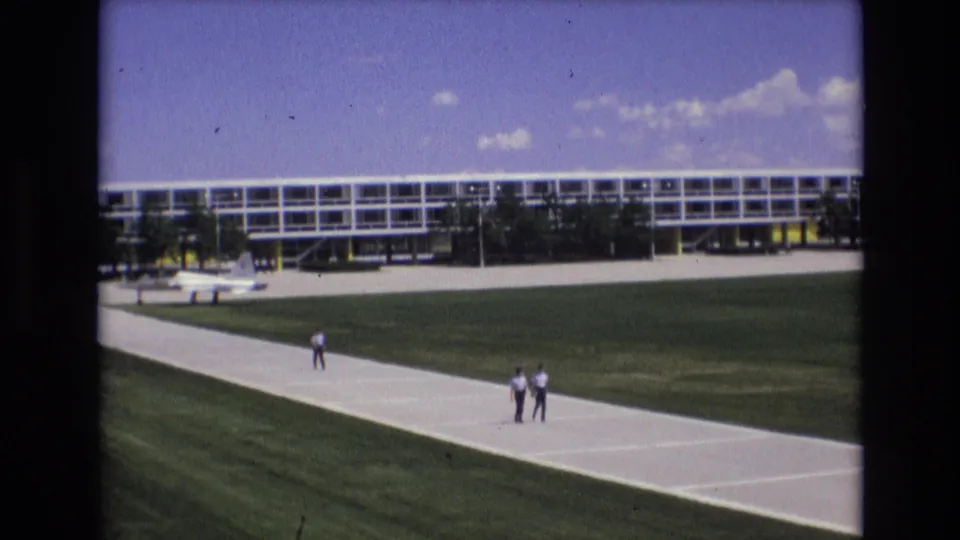 1973 Colorado Springs 8mm frame showing two distant men on a pale concrete walkway, with green grass, a long modern building, and blue sky behind them.