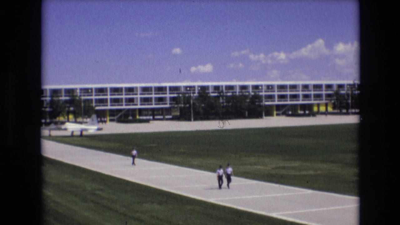 1973 Colorado Springs home-movie frame with two men walking side by side along a concrete path below a long windowed structure and open sky.